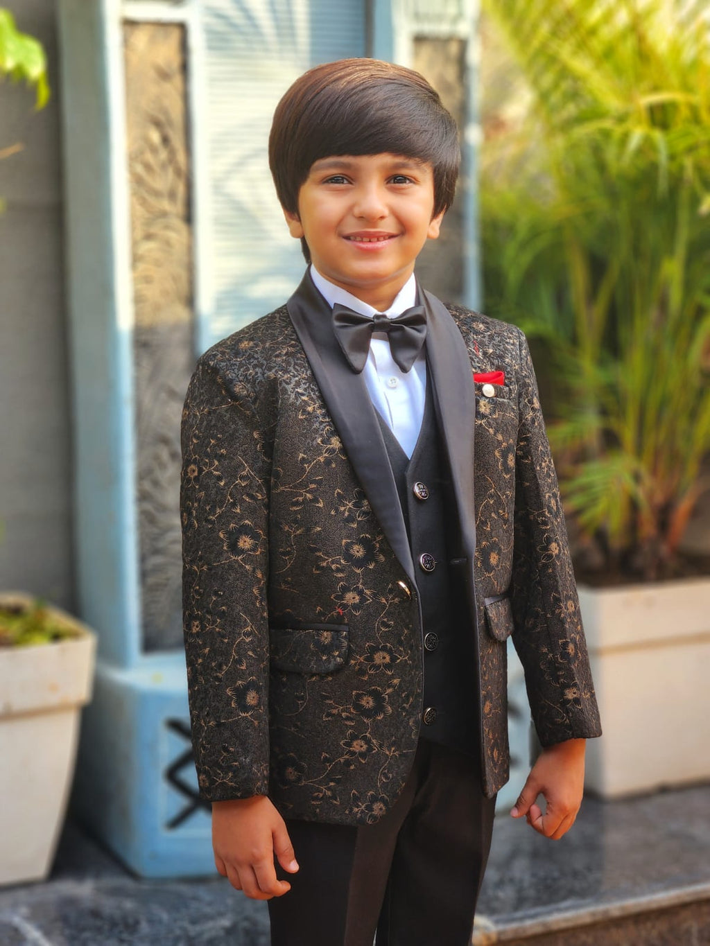 Young boy in a formal suit standing outdoors with plants in the background