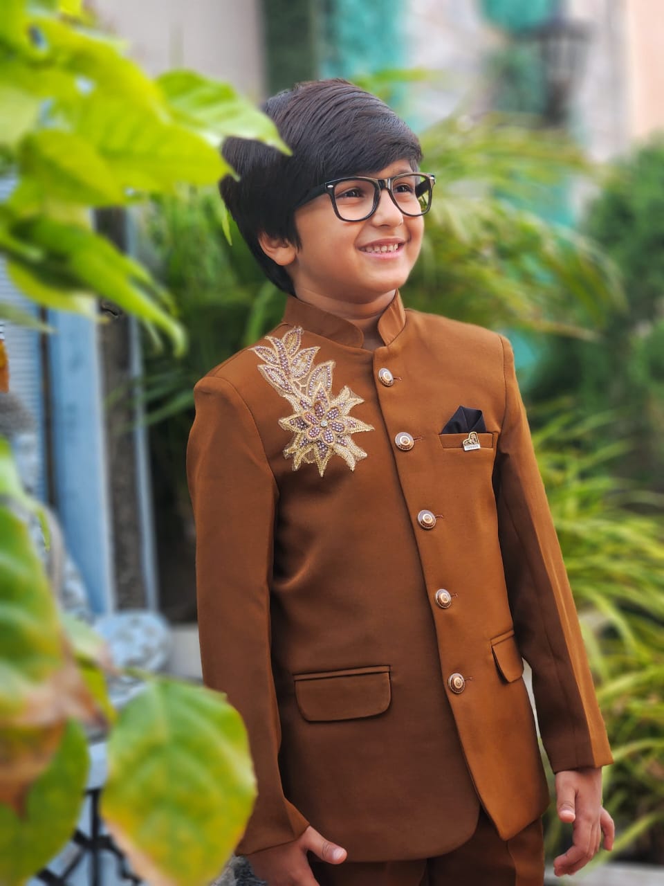 Child wearing a brown traditional outfit with decorative elements, standing outdoors with greenery.