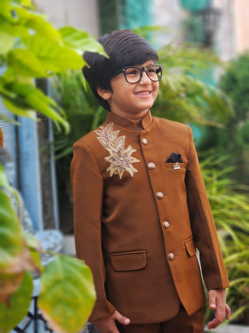 Child wearing a brown traditional outfit with decorative elements, standing outdoors with greenery.