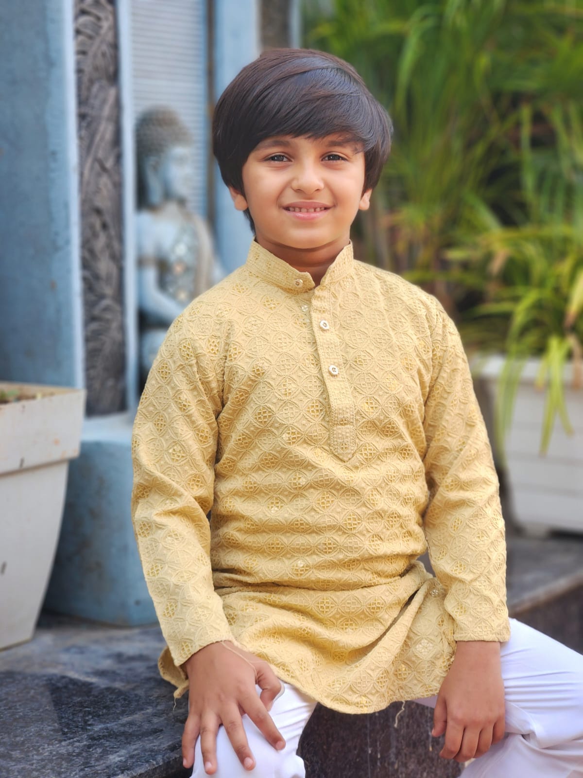 Child wearing a yellow kurta with white pants sitting outdoors.