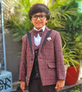 Young boy in a formal outfit with a maroon blazer and bow tie standing outdoors.