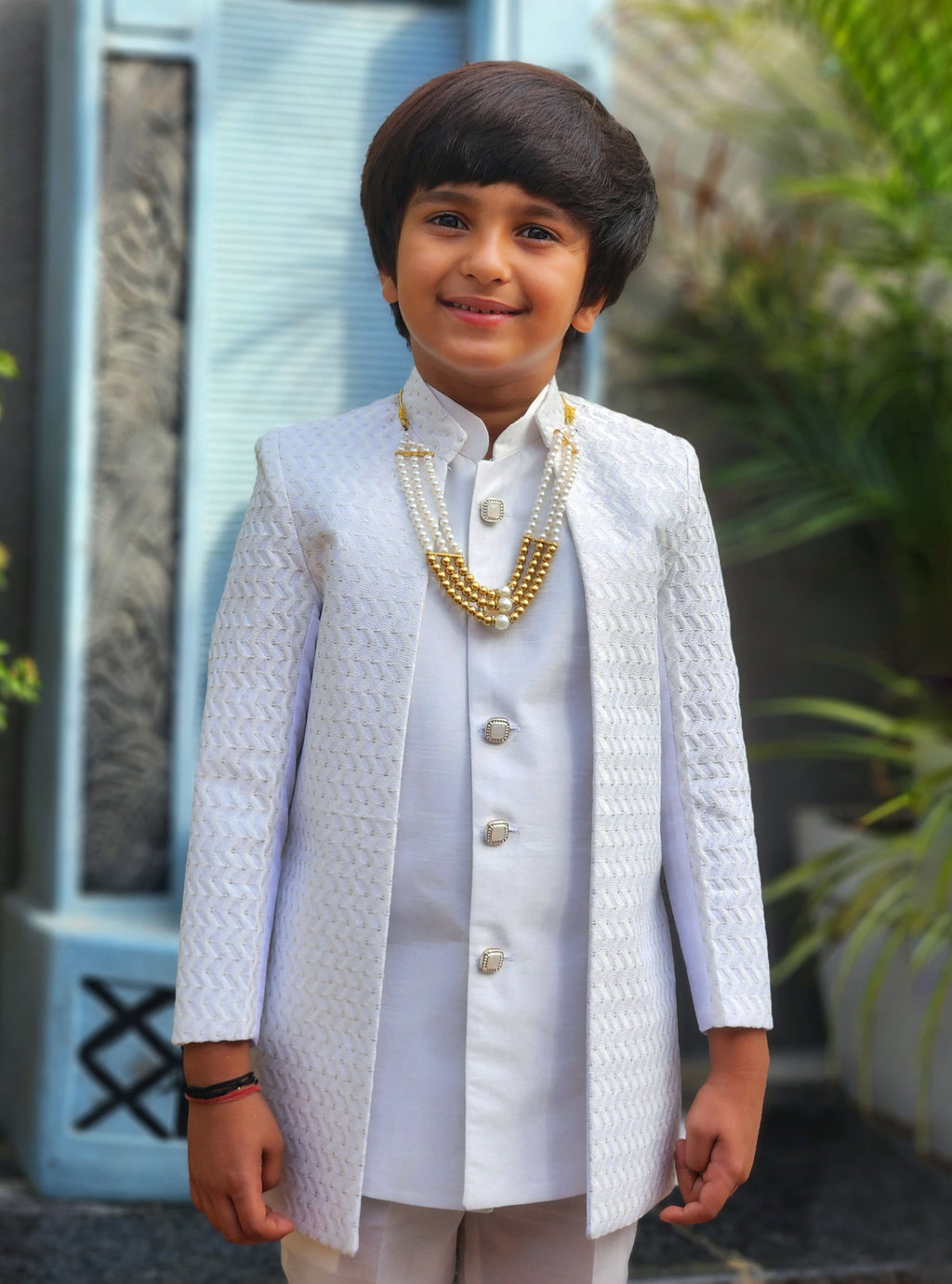 Young boy in a white traditional outfit with a necklace, standing outdoors.