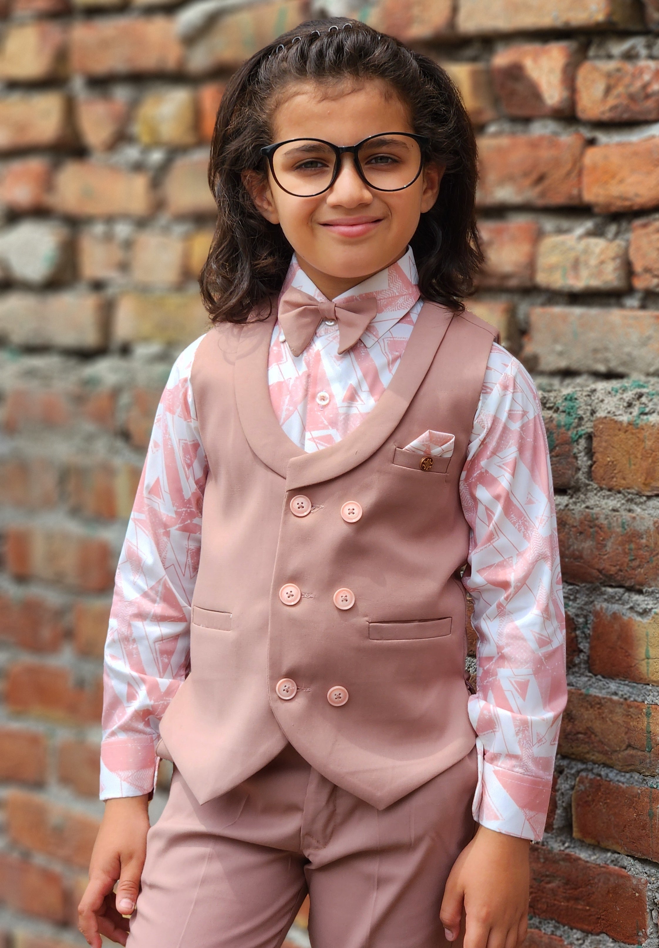 Child wearing a formal outfit with a vest and bow tie against a brick wall.