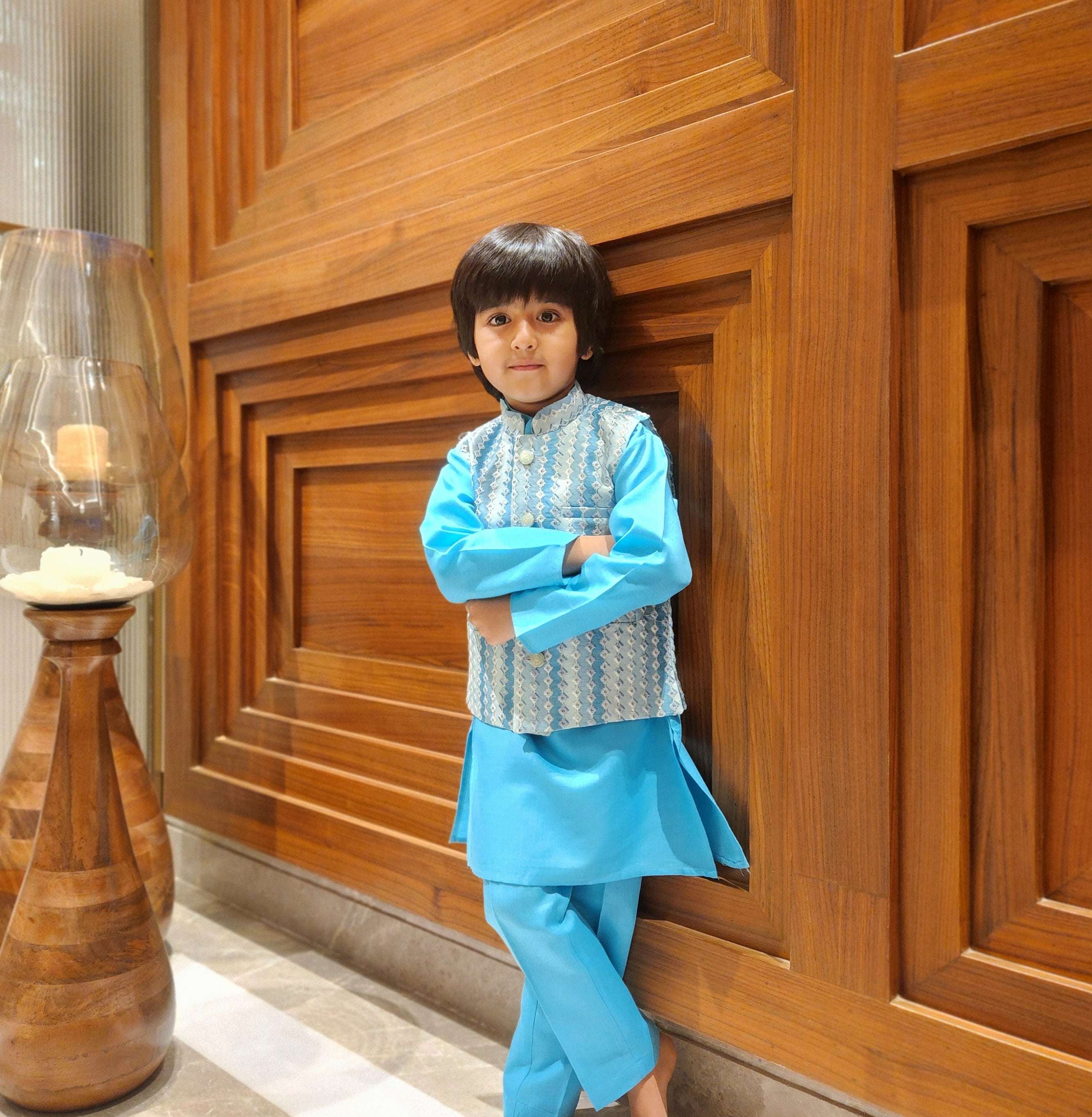 Child in a blue traditional outfit standing against a wooden wall.
