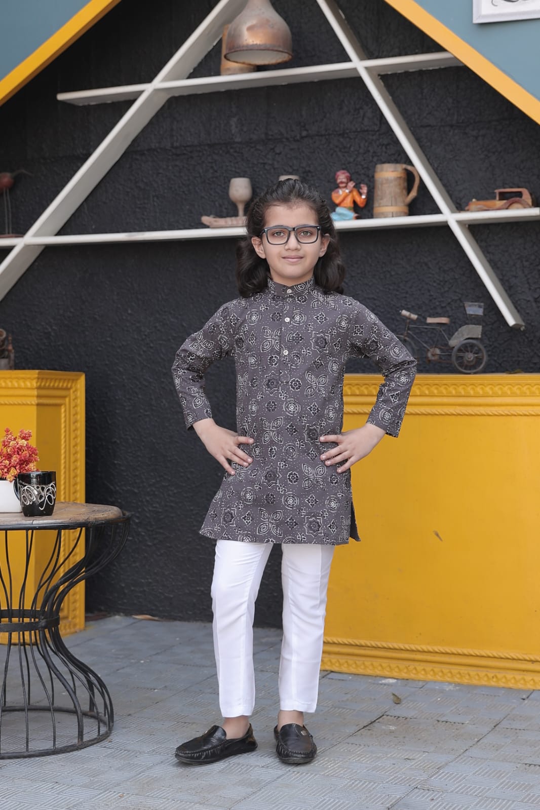 Child wearing a patterned kurta with white pants in front of a decorative wall.