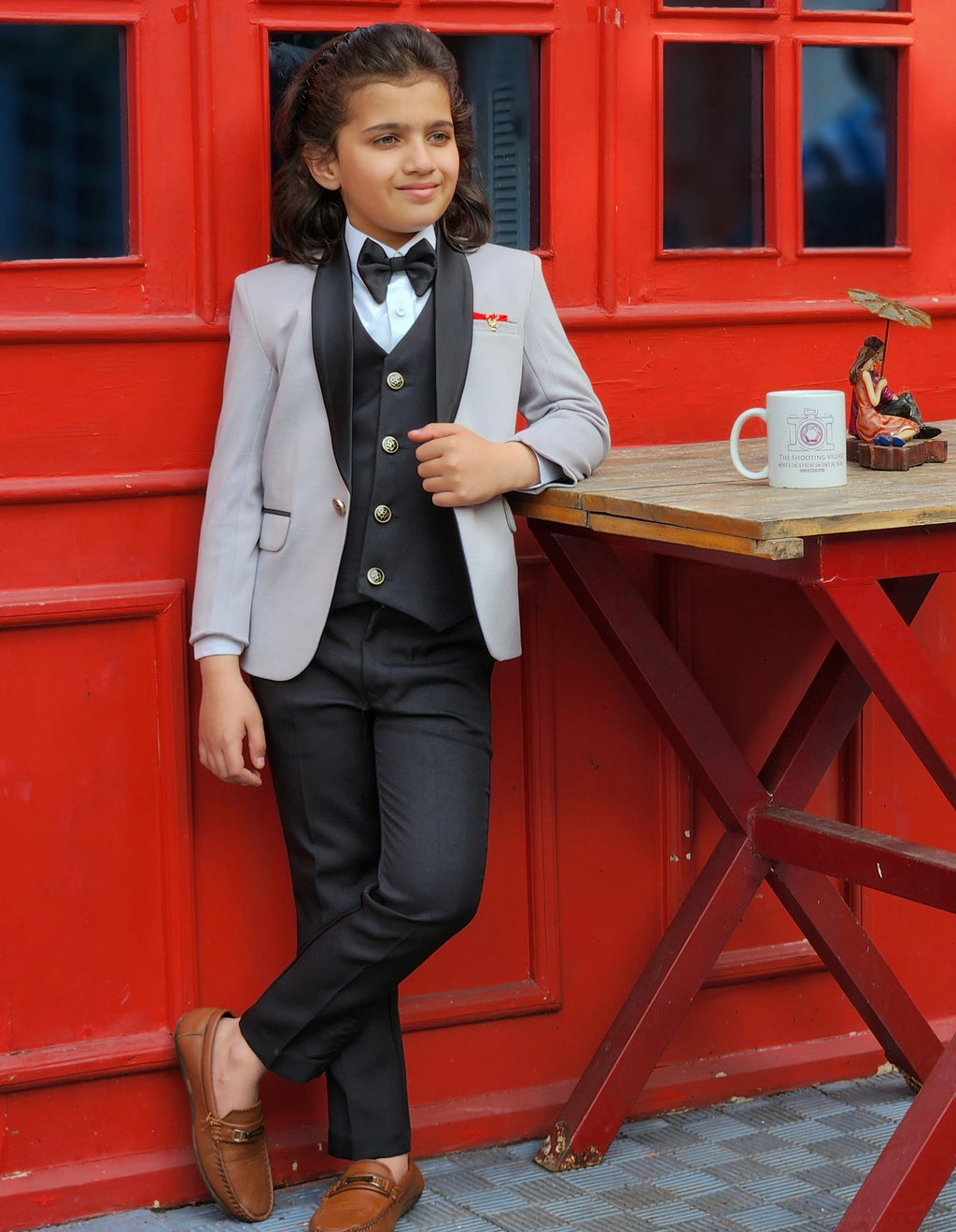 Child in formal attire leaning against a red wall with a table and mug.