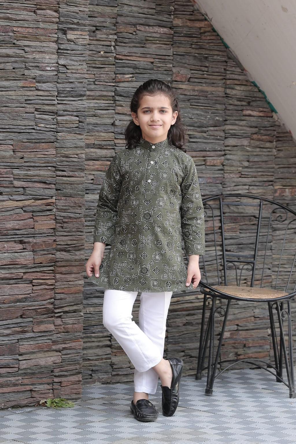 Child wearing a green kurta with white pants standing against a stone wall.