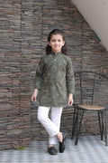 Child wearing a green kurta with white pants standing against a stone wall.