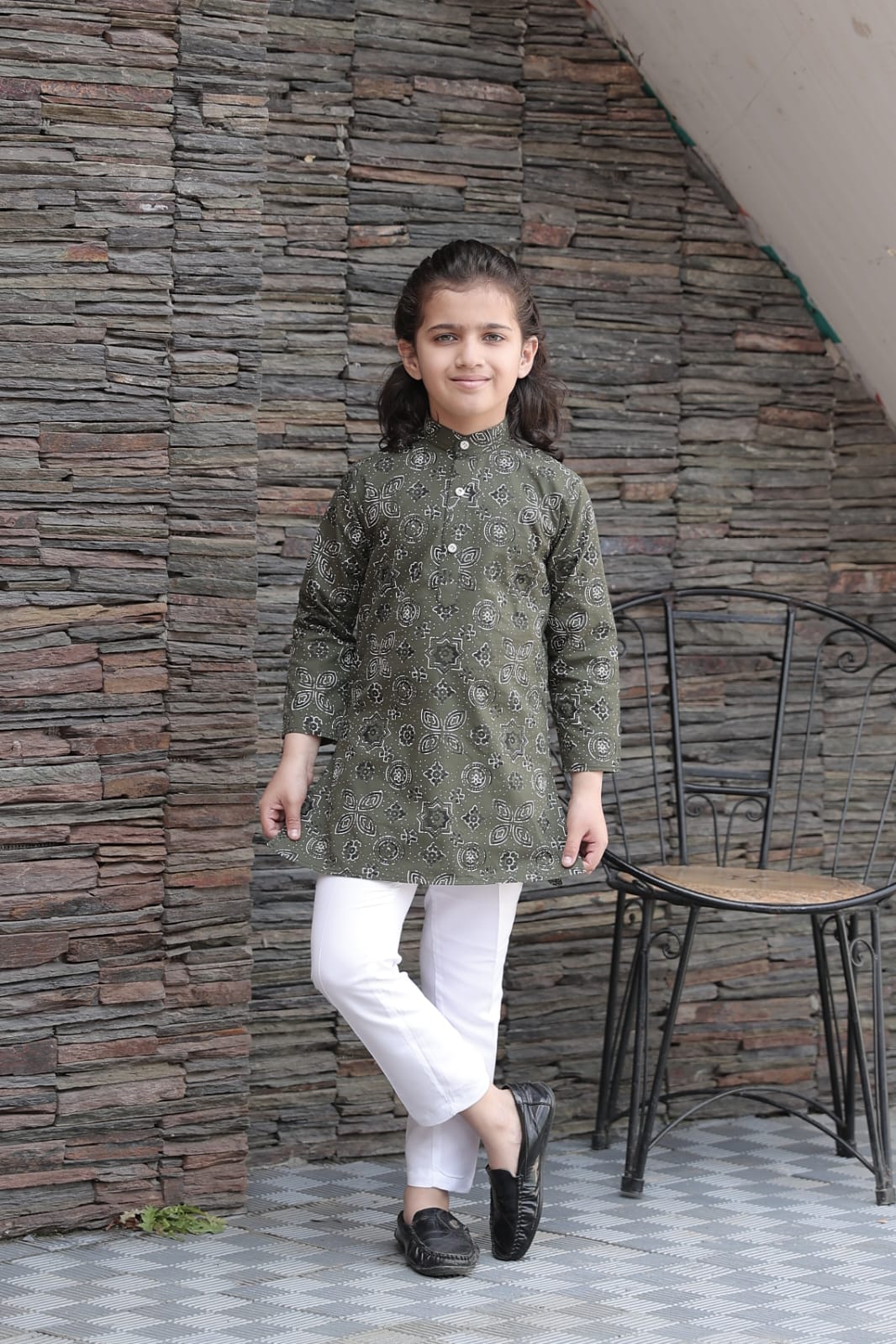 Child wearing a green kurta with white pants standing against a stone wall.