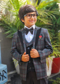 Child wearing a formal outfit with a plaid jacket and bow tie, standing outdoors with greenery in the background.