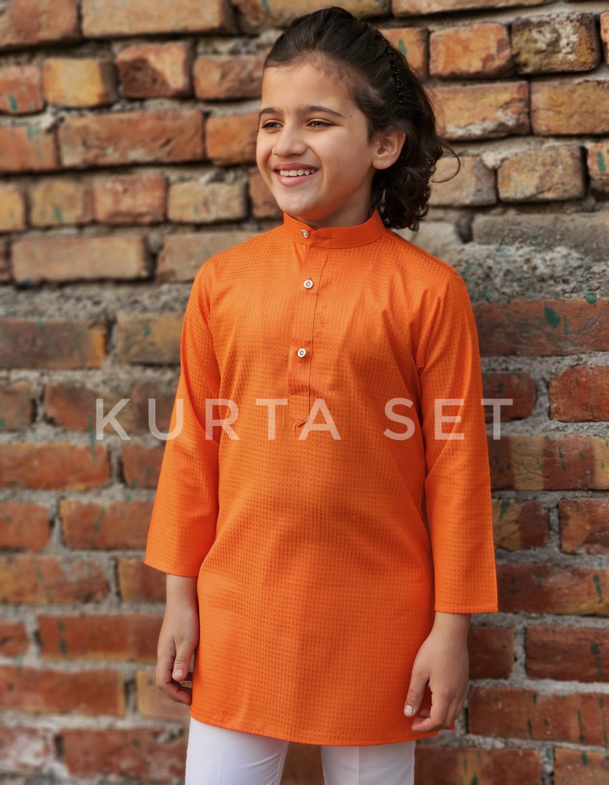 Child wearing an orange kurta set against a brick wall