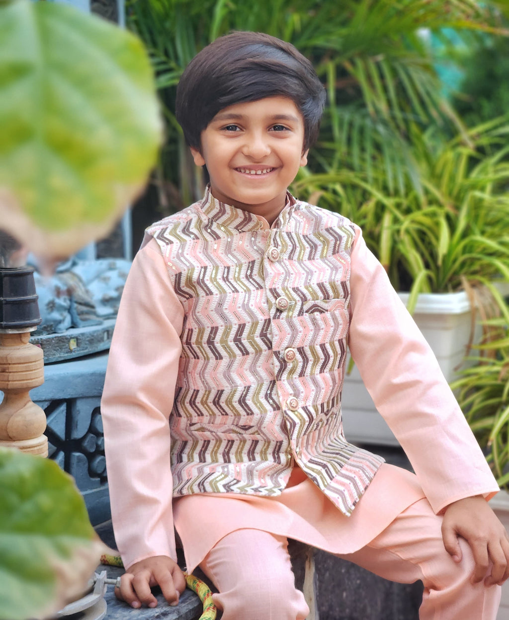 Child wearing a traditional outfit with a patterned vest and pink shirt, sitting outdoors.