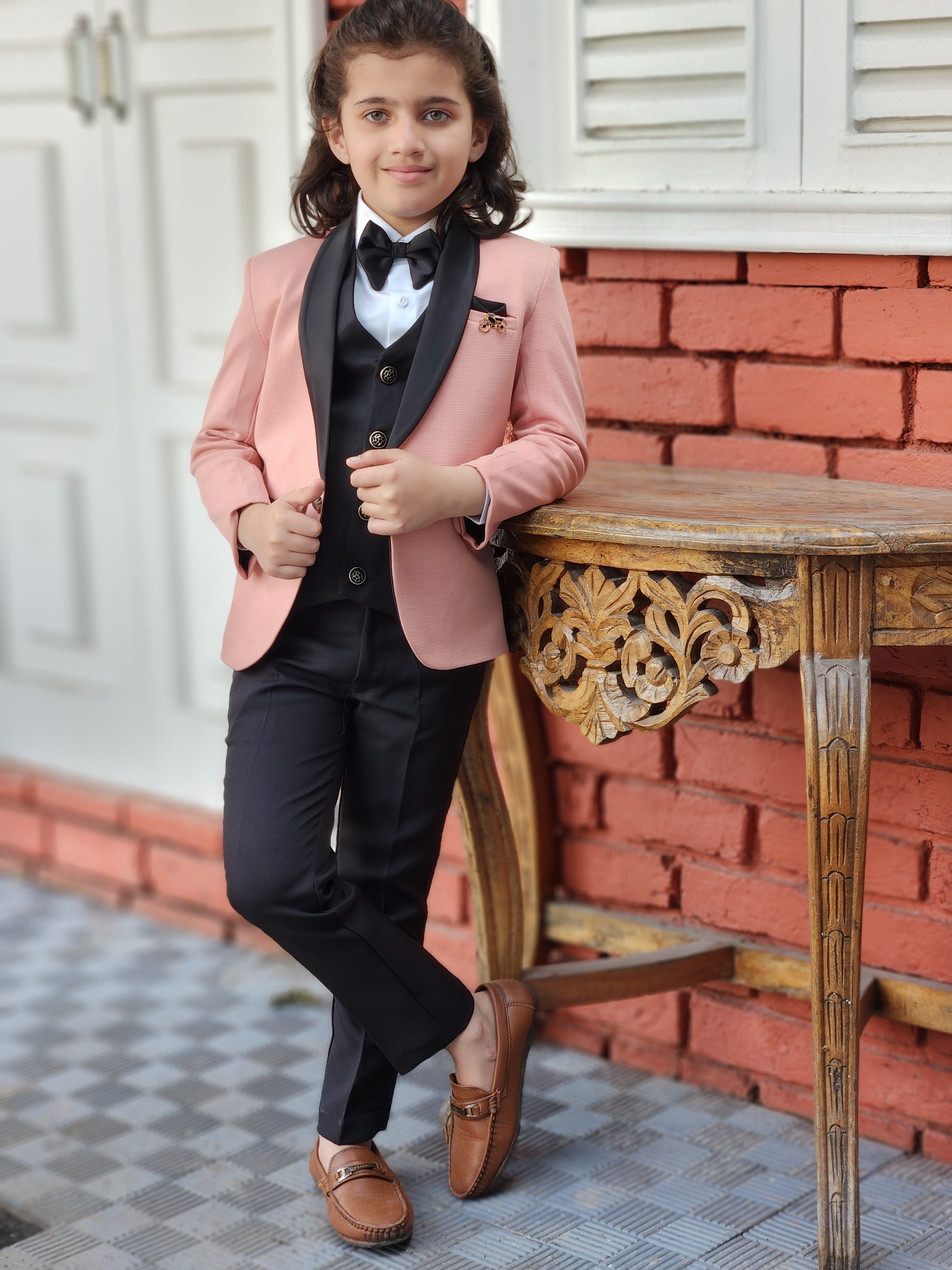 Person wearing a pink blazer and black dress over a white shirt with a black tie, standing in a room with red brick walls and a decorative chair.