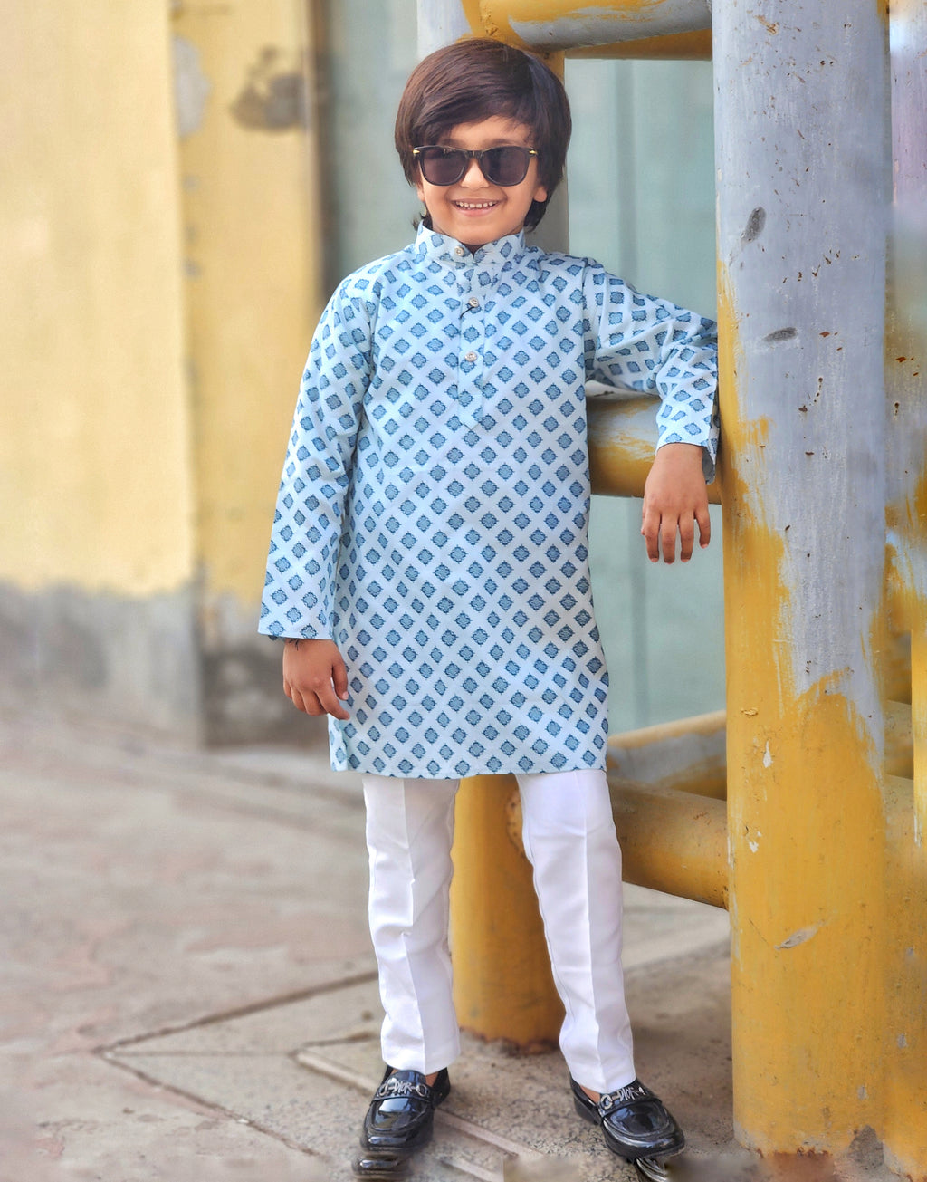 Child wearing a blue and white patterned kurta with white pants, standing against a yellow wall.