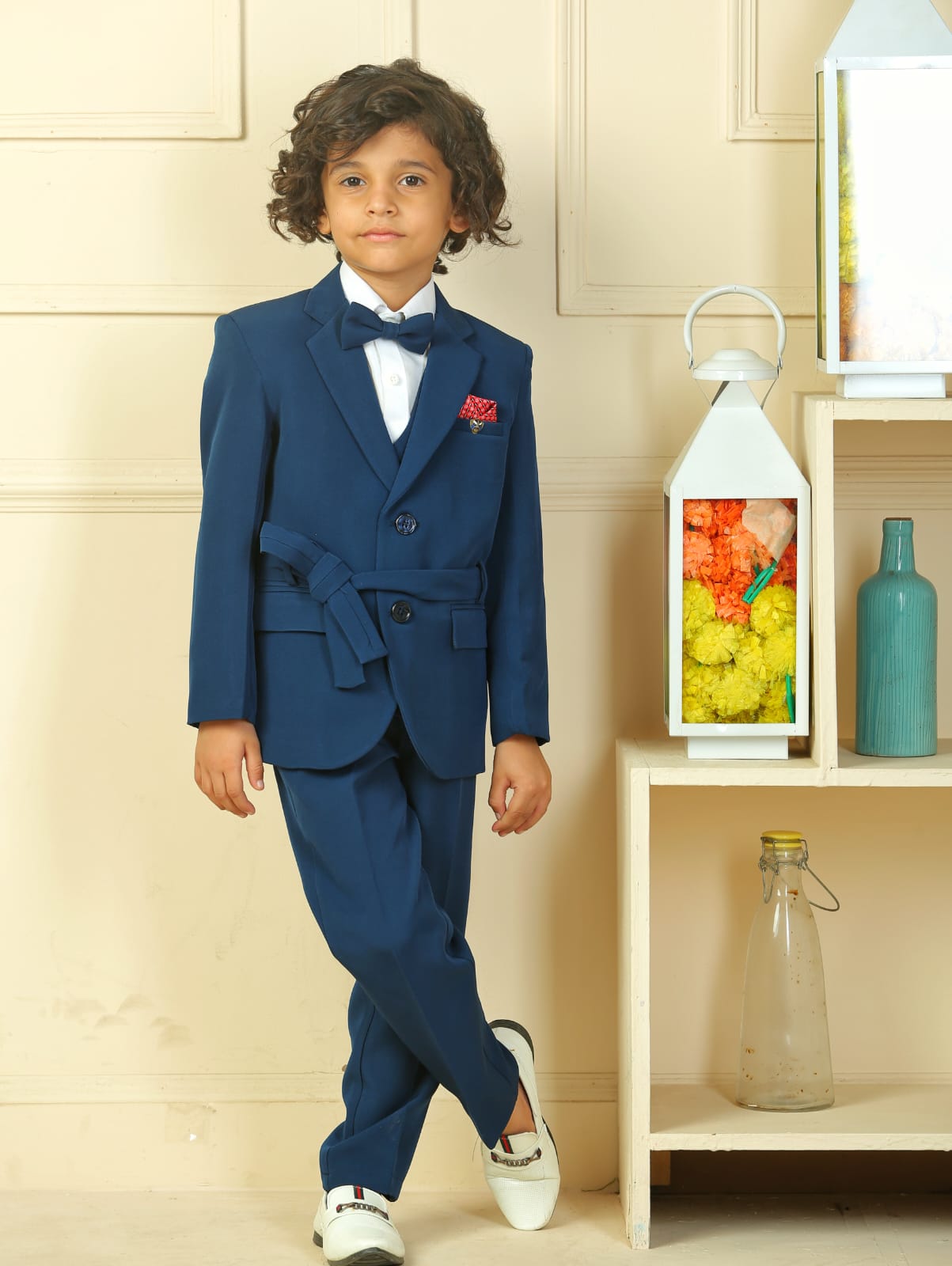 Child wearing a blue suit with a white shirt and bow tie, standing in a room with a shelf in the background.