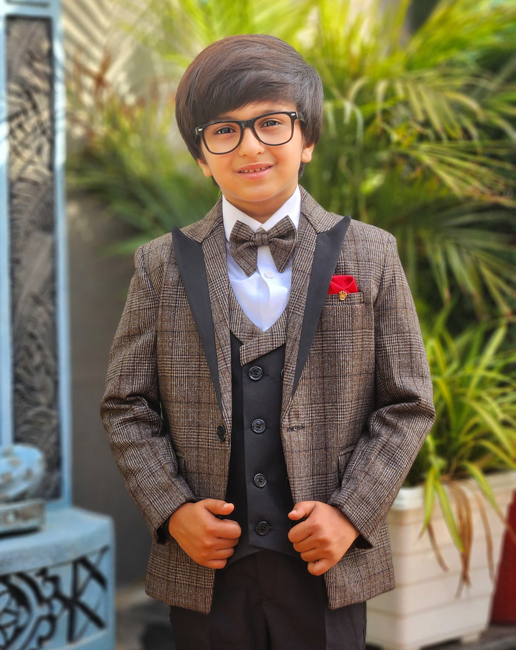 Young boy in a formal suit with a bow tie and red flower, standing outdoors.