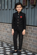 Young boy in a black suit with a red pocket square standing against a brick wall.